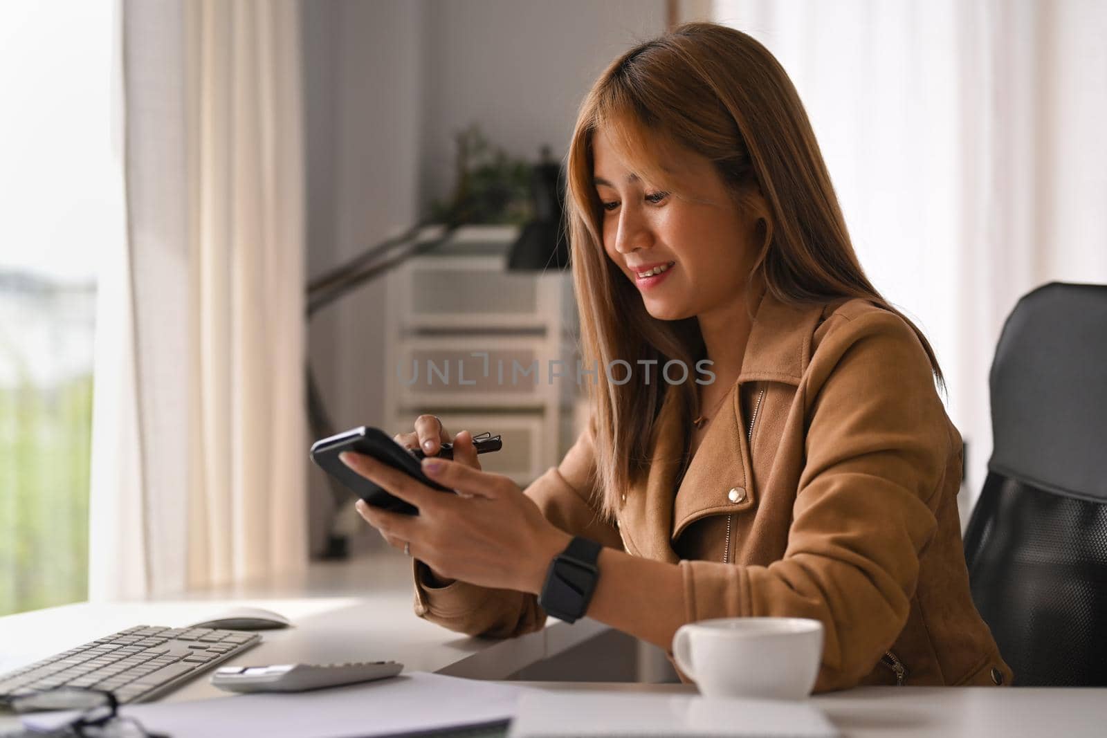 Smiling young creative woman sitting in bright office and using smart phone.