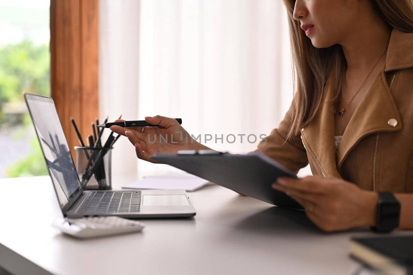 Cropped shot of female investor holding clipboard and pointing on laptop computer screen by prathanchorruangsak