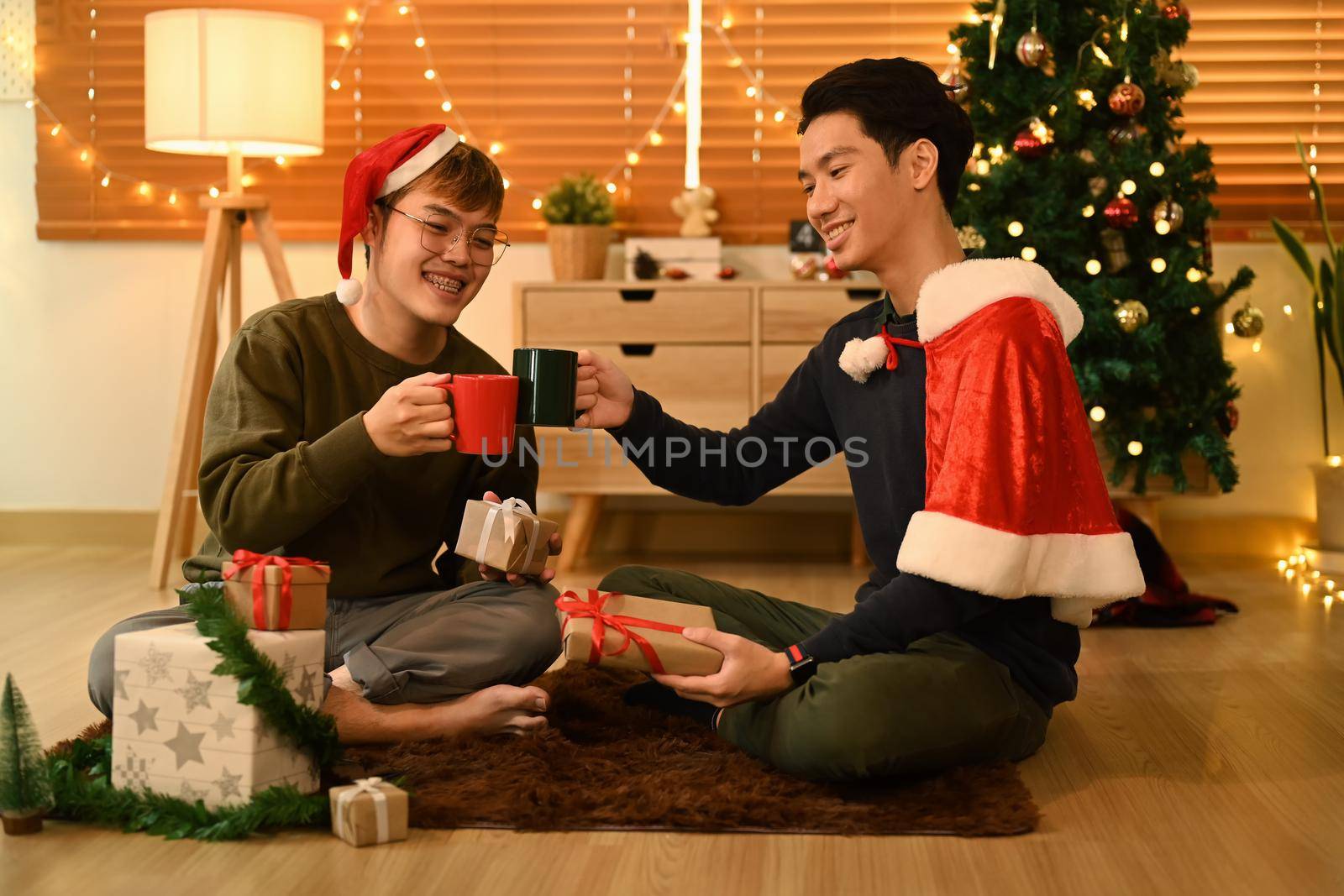 Two happy man next to a decorated Christmas tree and enjoying drinking hot chocolate together.