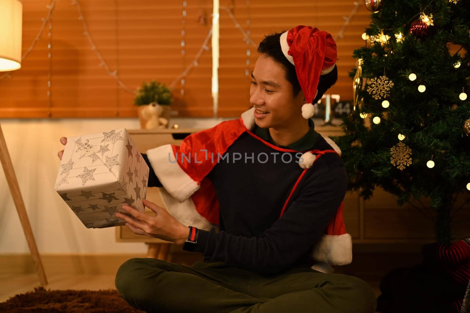 Smiling man in Santa hat opening Christmas gifts and sitting in decorated room for celebrating New year and Christmas festive by prathanchorruangsak