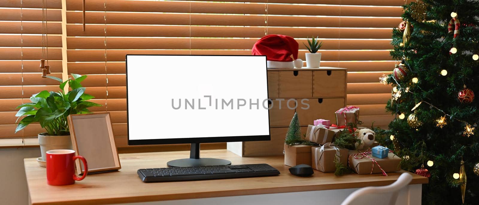 Computer with empty display, coffee cup and gift boxes on wooden table near Christmas tree.