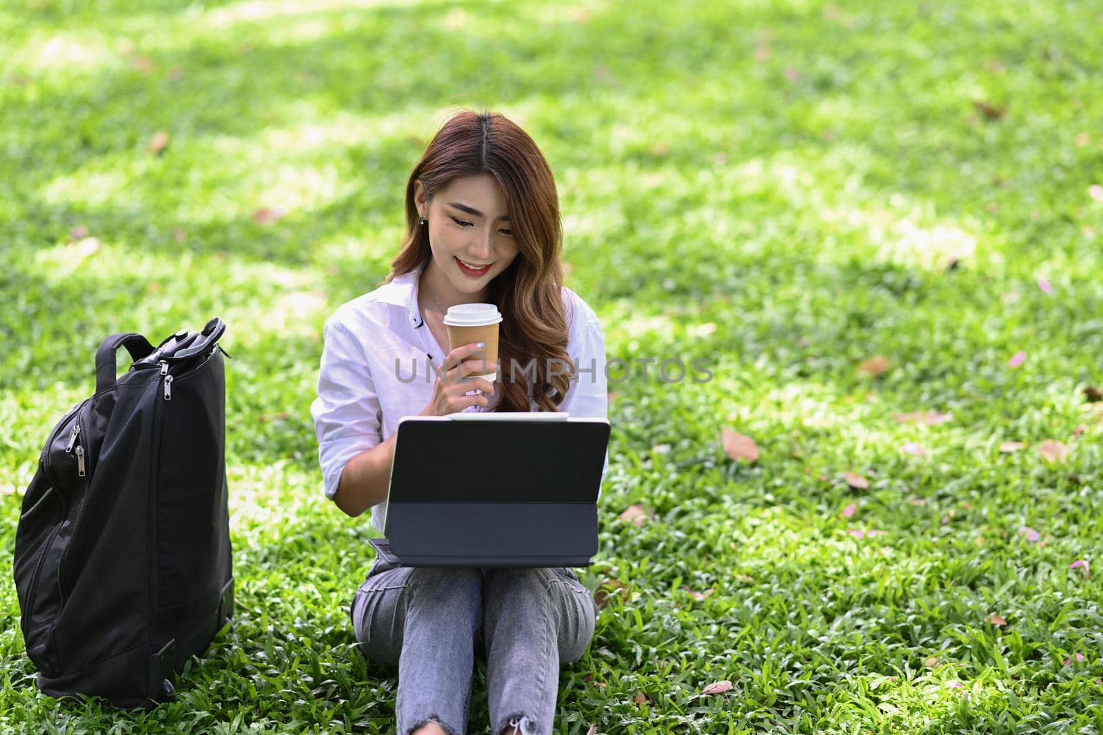 Beautiful asian woman drinking coffee and working with computer tablet in the park. by prathanchorruangsak