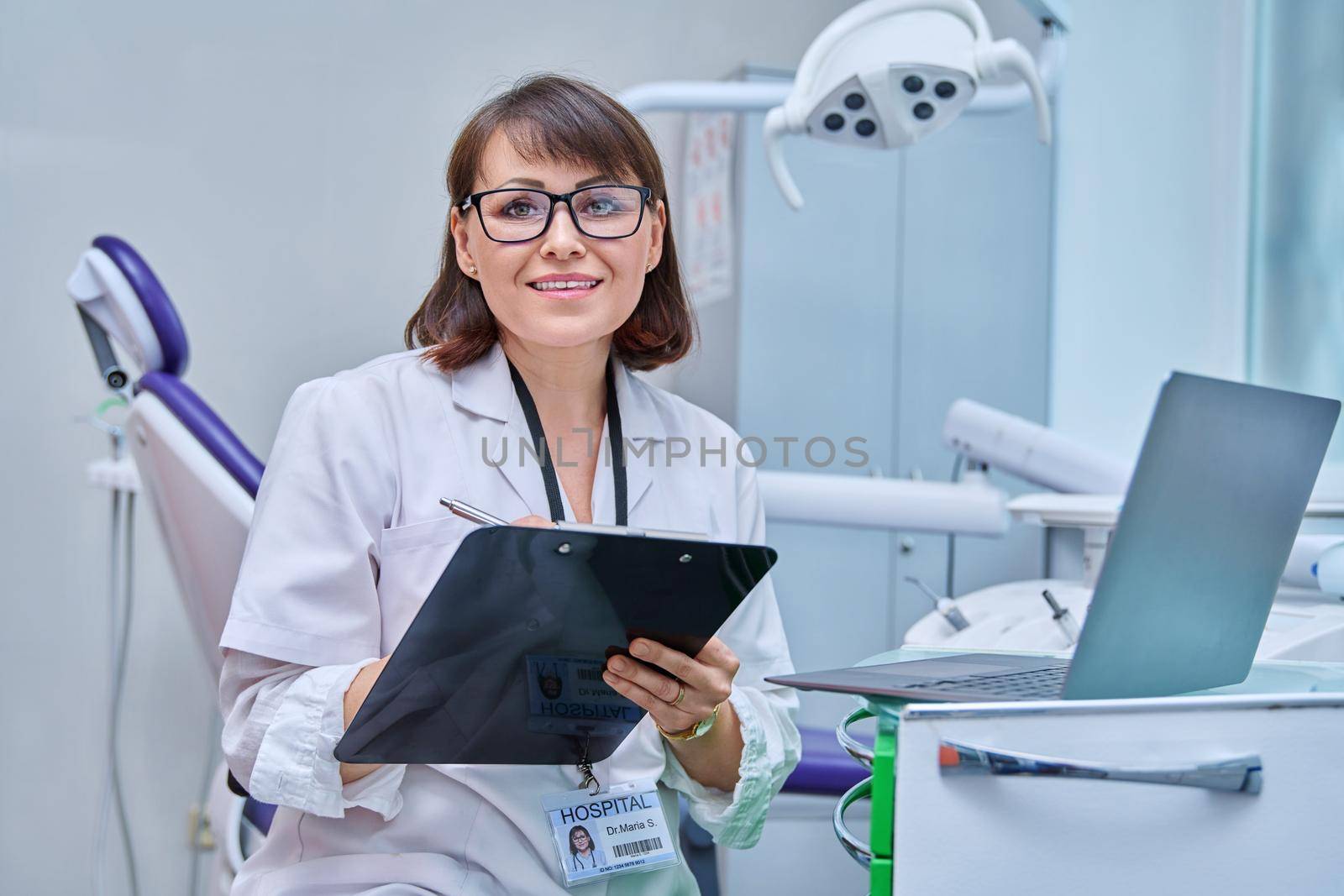 Doctor dentist working in office, looking at camera, using laptop, making notes on clipboard. Dentistry, medicine, hygiene, treatment, dental health care concept