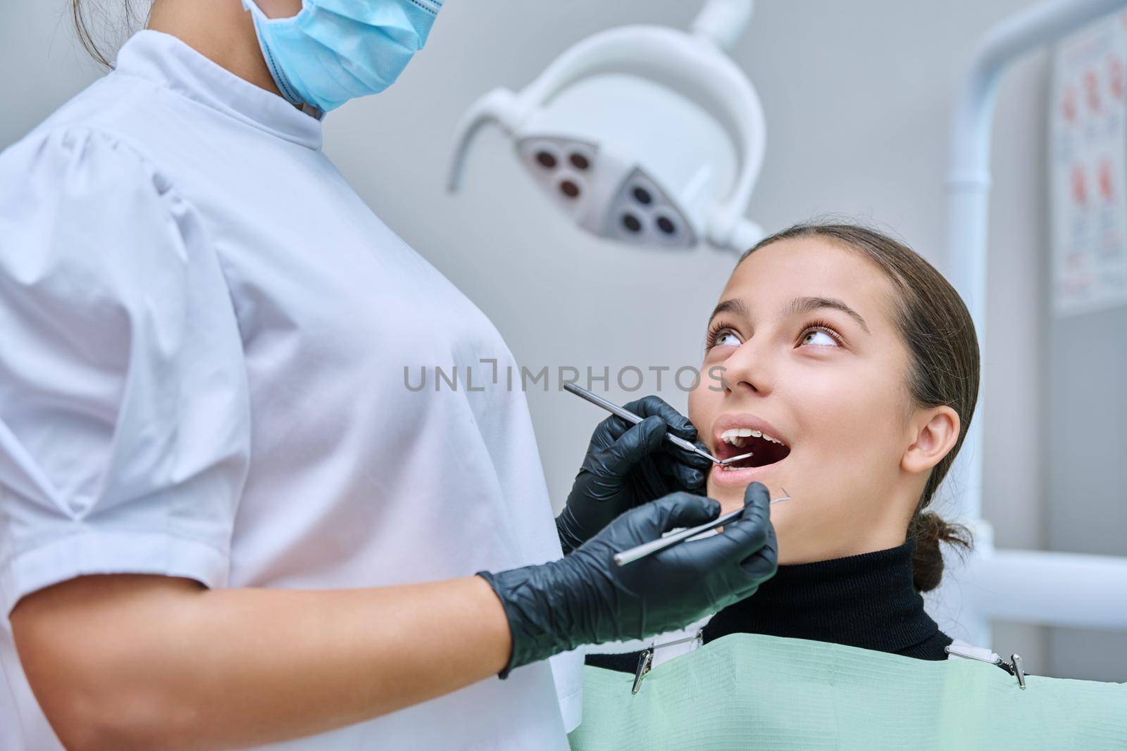 Young teenage female at dental checkup in clinic. Teenage girl sitting in chair, doctor dentist with tools examining patient's teeth. Adolescence, hygiene, dentistry, treatment, dental health care