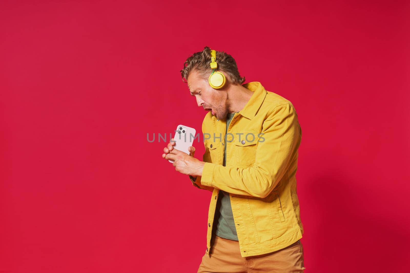 Joyful man sing while recording his voice using smartphone. Singing handsome man enjoying his favorite song using phone and wireless headphones wearing jeans yellow jacket isolated on red background.