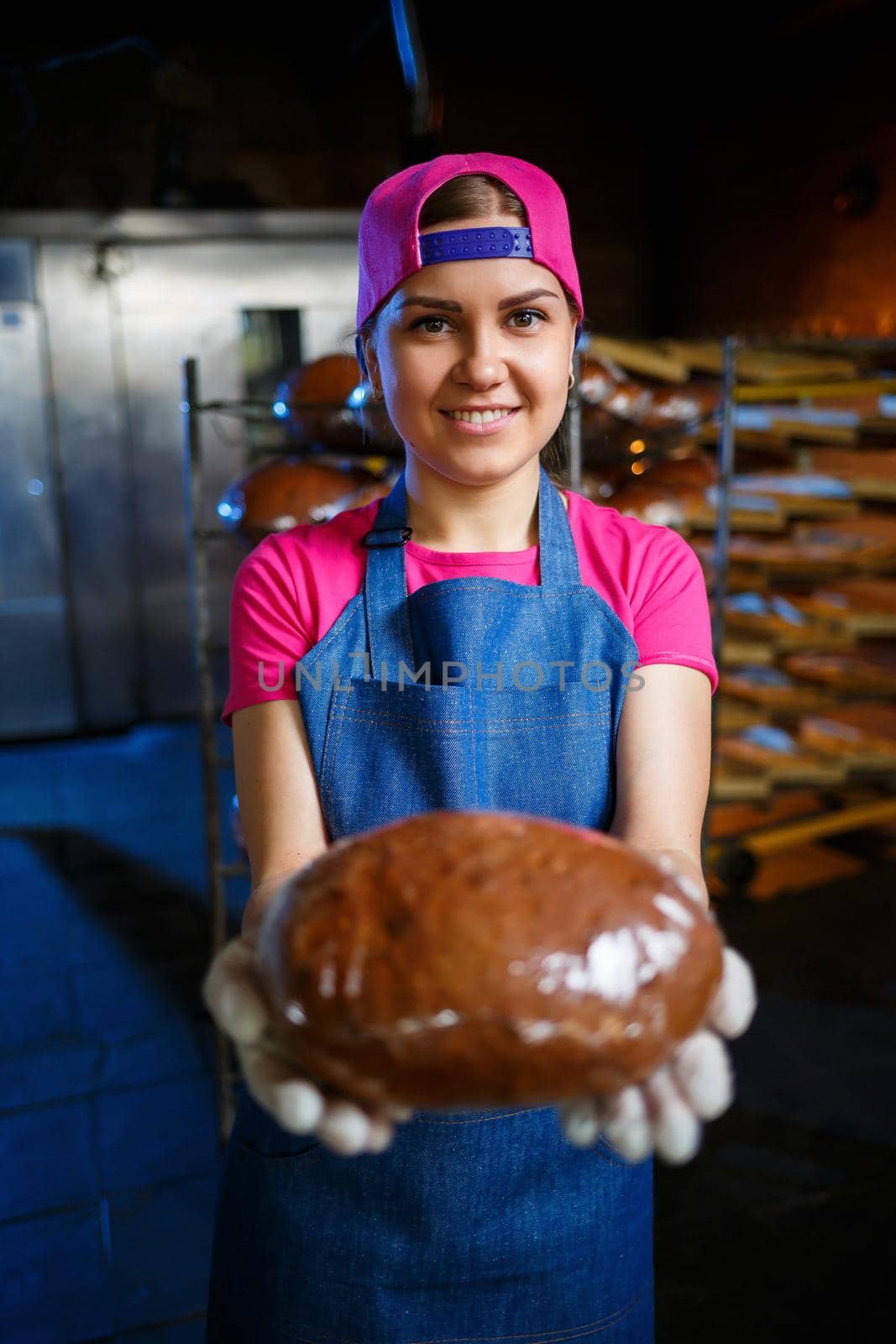 A baker girl takes hot bread in a bakery against the background of shelving with bread. Industrial production of bread. The stage of baking in a bakery