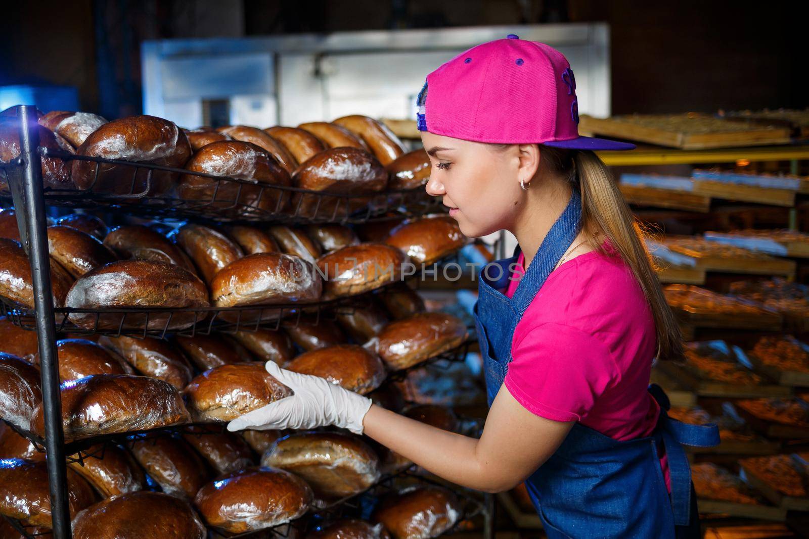 A young girl works in a bakery. She puts bread on a shelf. Woman baker at workplace in a bakery. A professional baker holds bread in his hands. Bread production concept