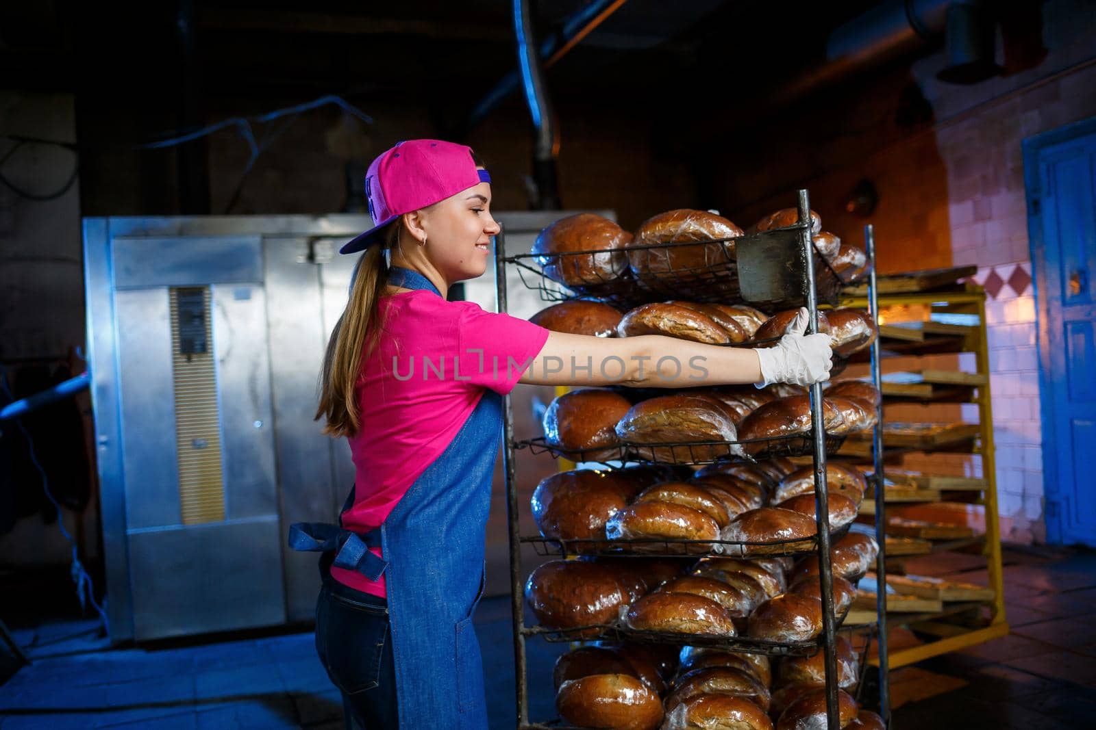 Professional baker - a young, pretty woman in a jeans apron holds fresh bread against the background of a bakery or bakery. Bakery products. Bread production