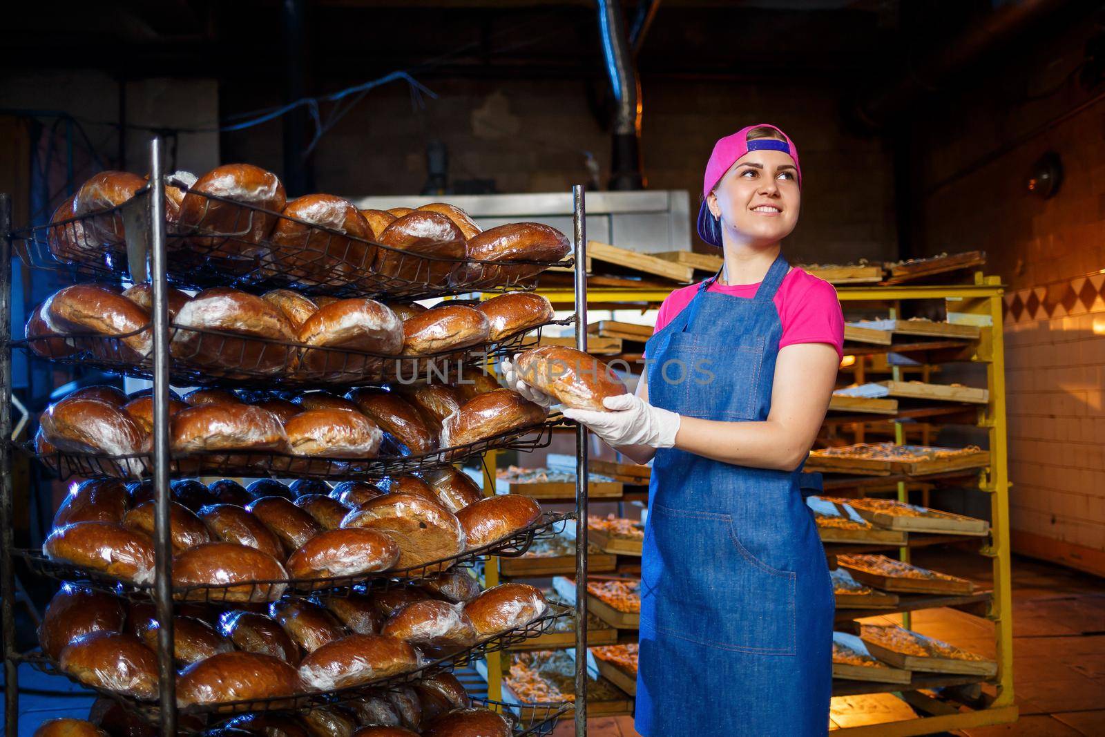 Professional baker - a young, pretty woman in a jeans apron holds fresh bread against the background of a bakery or bakery. Bakery products. Bread production