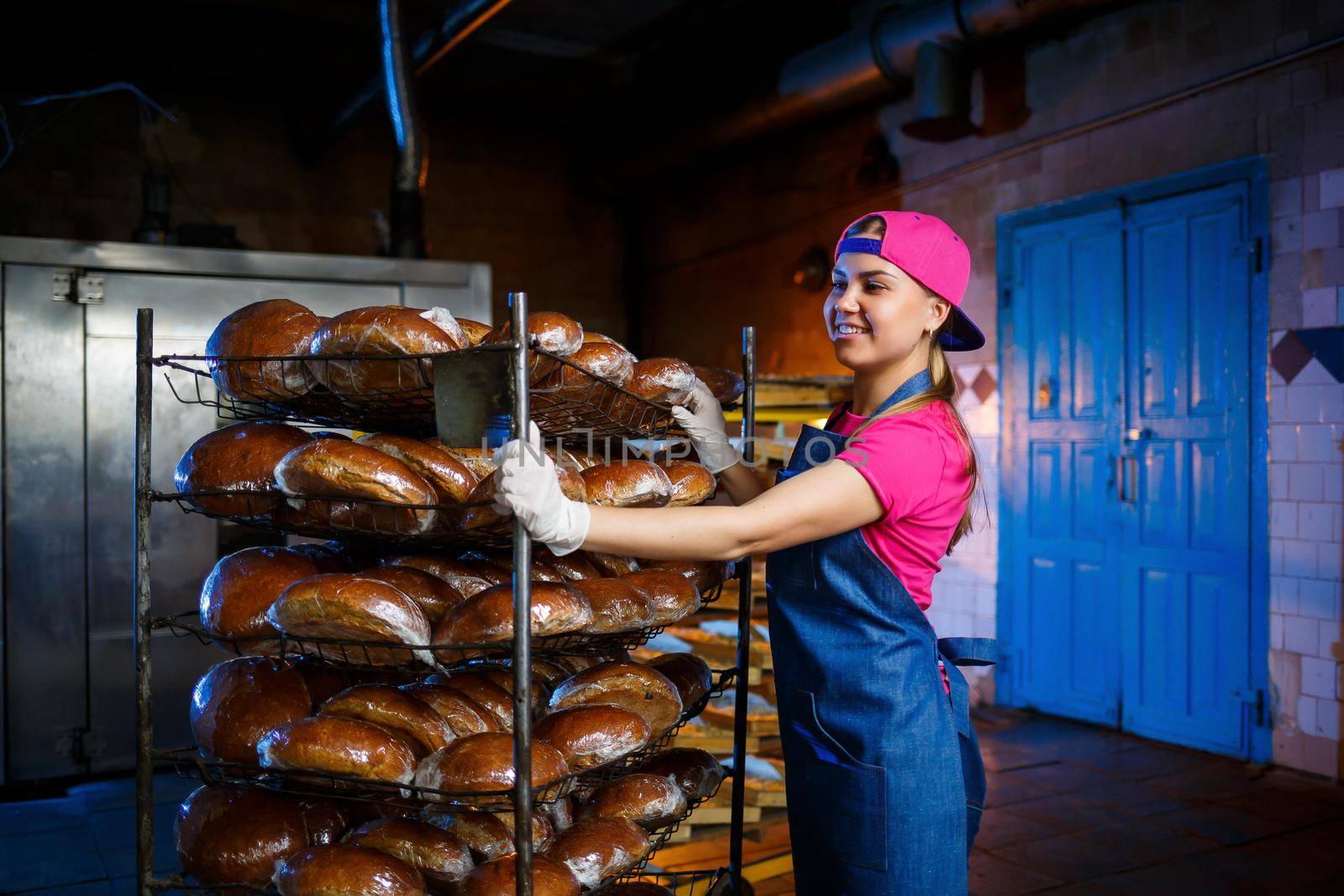 A baker girl takes hot bread in a bakery against the background of shelving with bread. Industrial production of bread. The stage of baking in a bakery