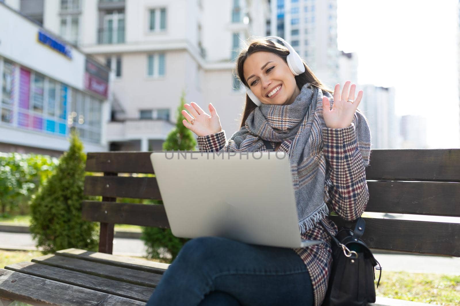 young woman in headphones with a laptop communicates via video link while sitting on the street with a smile on her face.