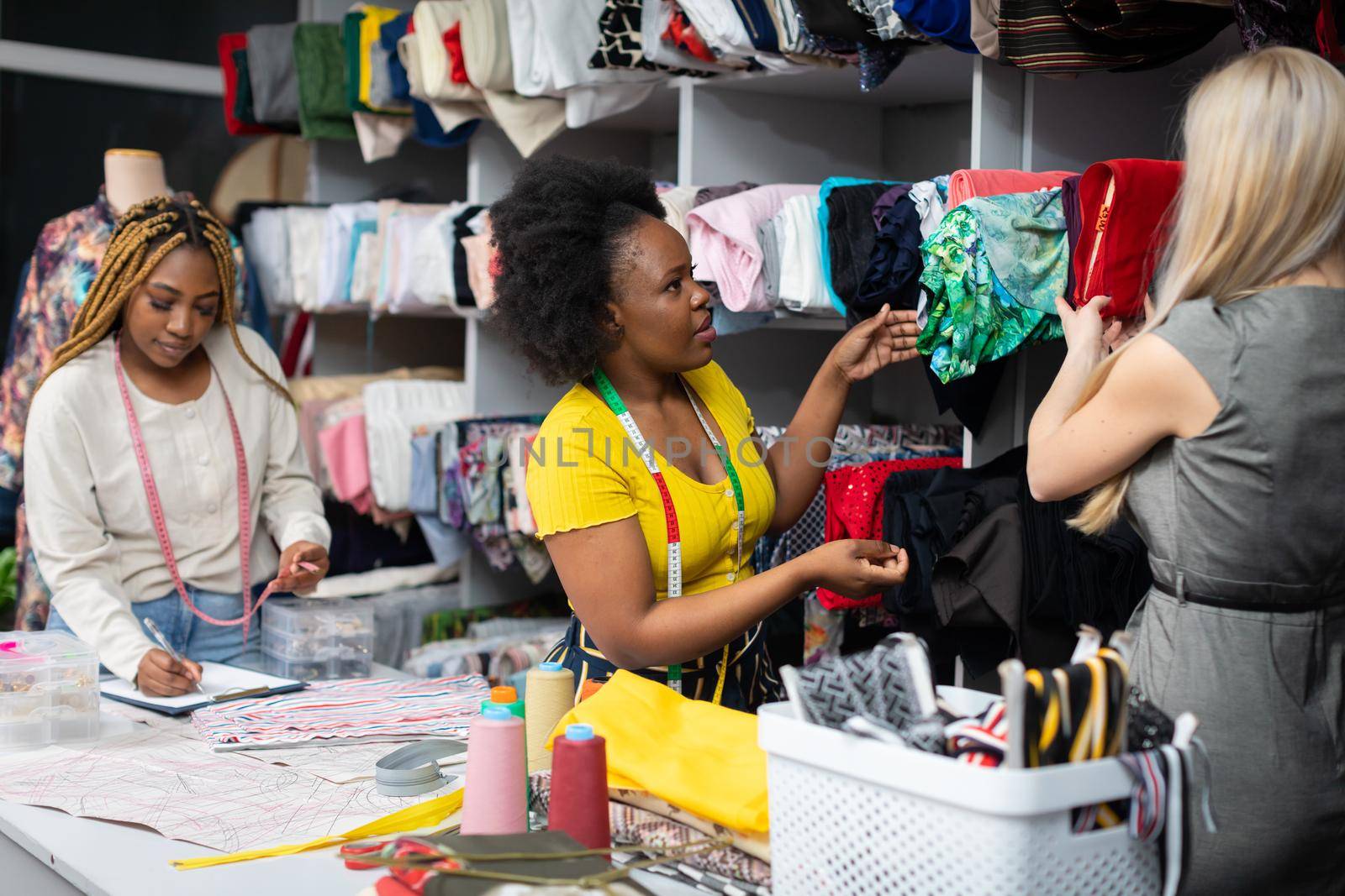 A dark-skinned shopkeeper shows a blonde woman what materials are most appropriate for a summer dress. Two African women and a European woman work in a tailor's shop.