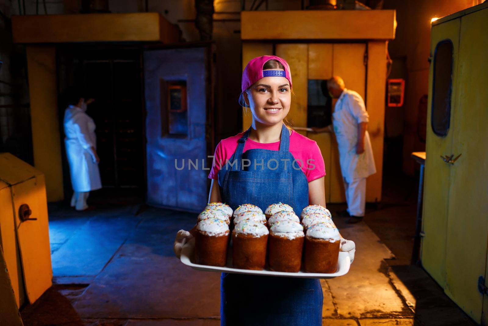 Girl baker holds a tray with hot pastries in the bakery. She is wearing a denim frock and a cap. Production of bakery products. Rack with fresh crispy pastries.