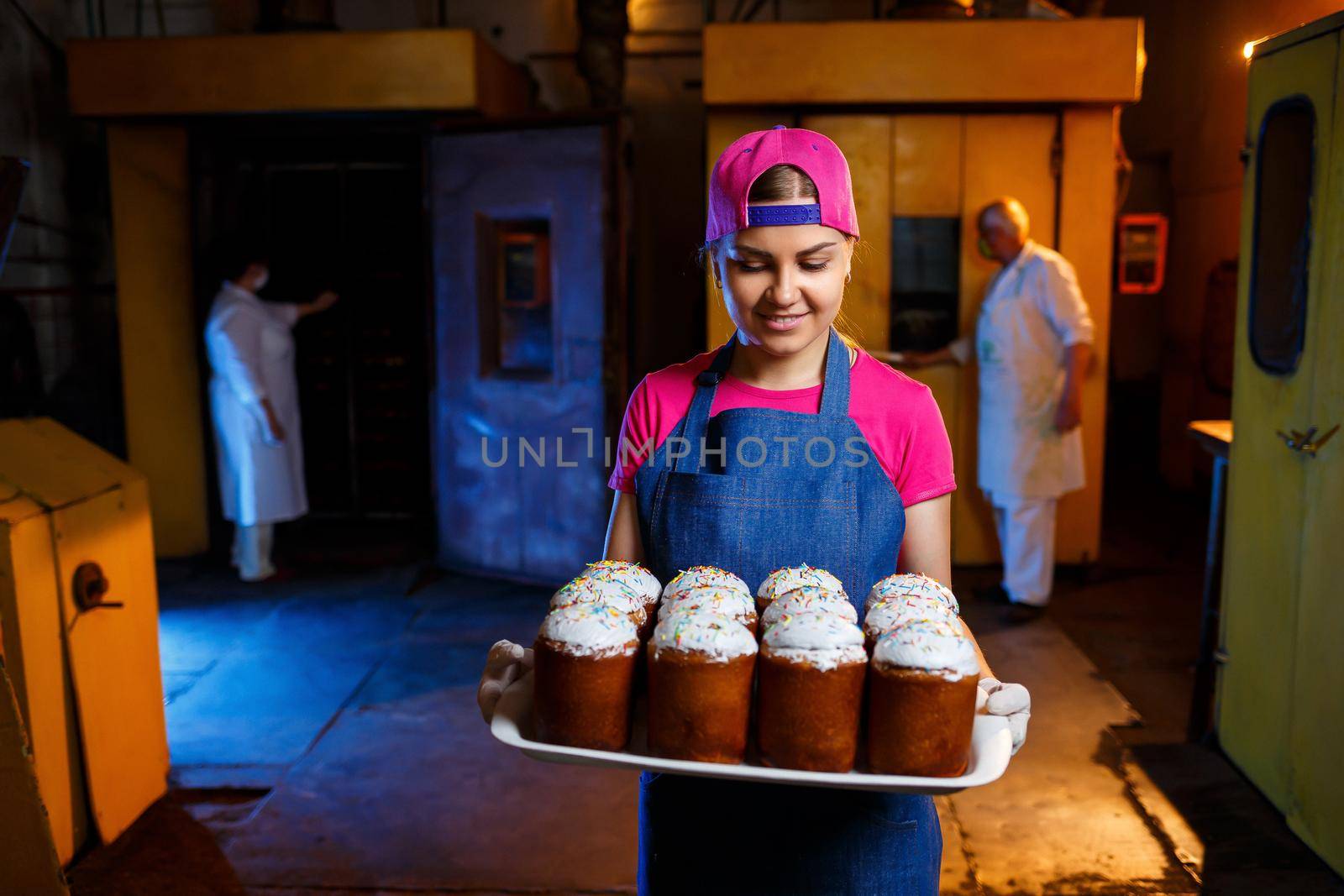 Girl baker holds a tray with hot pastries in the bakery. She is wearing a denim frock and a cap. Production of bakery products. Rack with fresh crispy pastries.