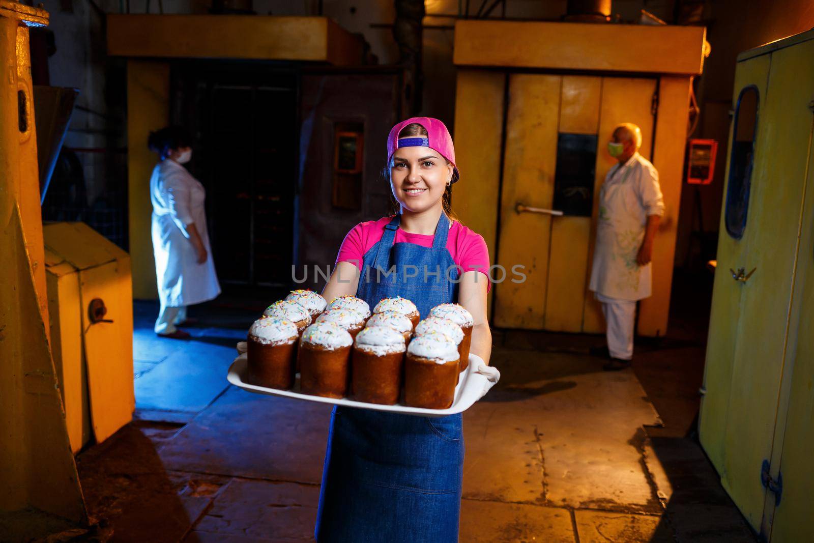 Girl baker holds a tray with hot pastries in the bakery. She is wearing a denim frock and a cap. Production of bakery products. Rack with fresh crispy pastries.