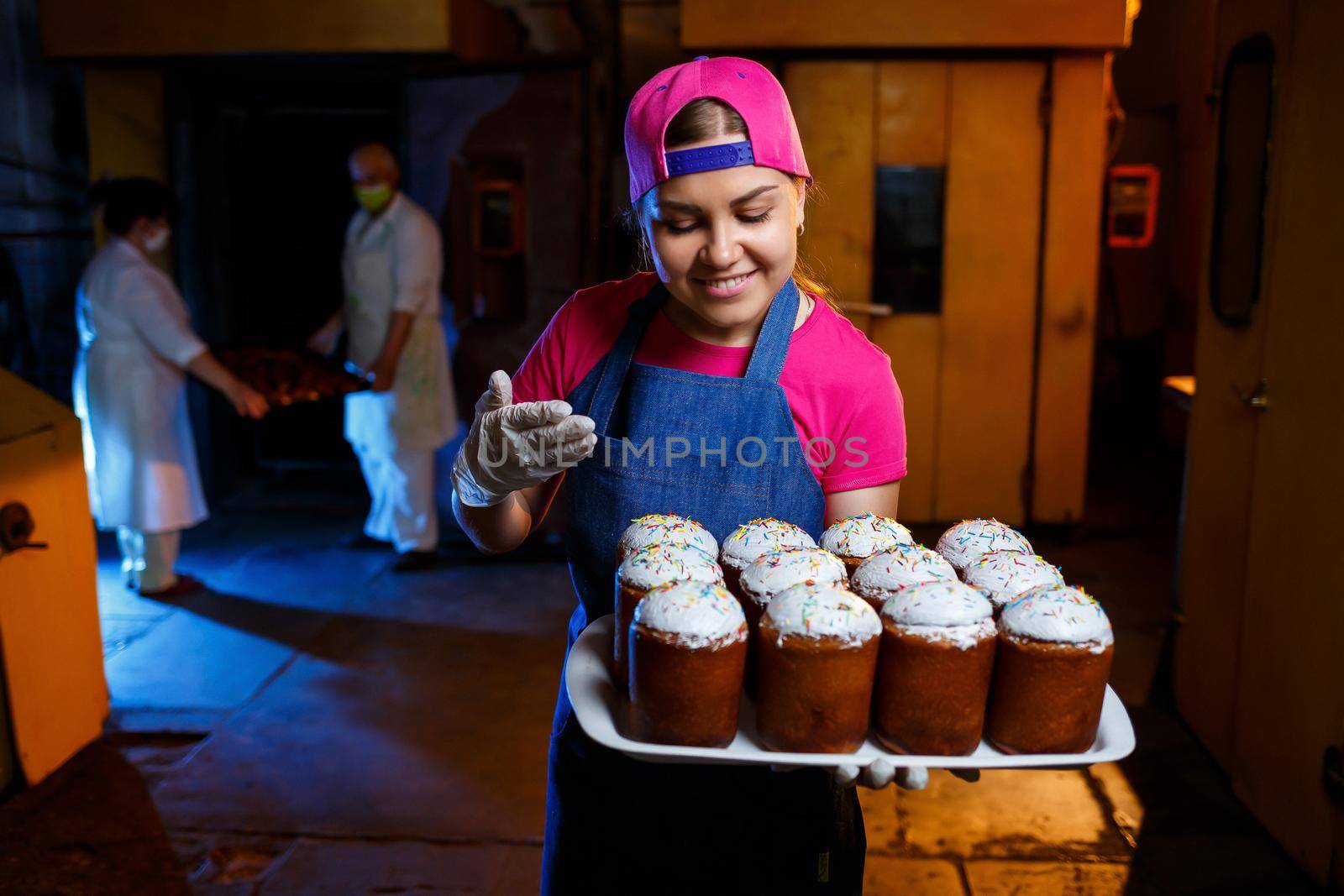 Girl baker holds a tray with hot pastries in the bakery. She is wearing a denim frock and a cap. Production of bakery products. Rack with fresh crispy pastries.