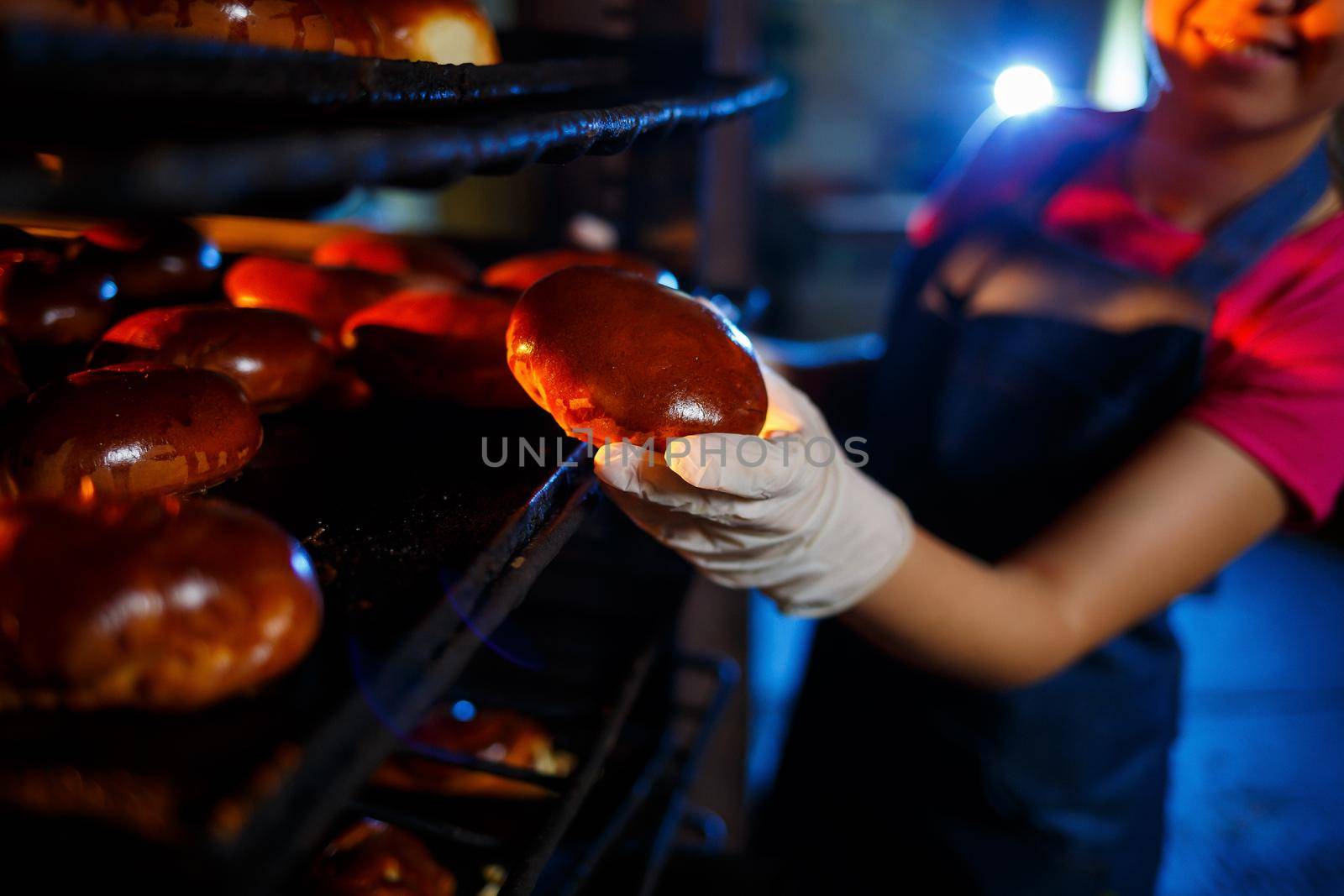 Girl baker holds fresh pies in the hands of the bakery. She is wearing a denim frock and a cap. Production of bakery products. Rack with hot crispy pastries.