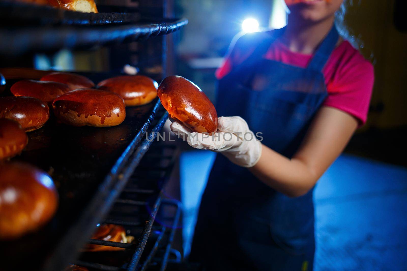 Girl baker holds fresh pies in the hands of the bakery. She is wearing a denim frock and a cap. Production of bakery products. Rack with hot crispy pastries.