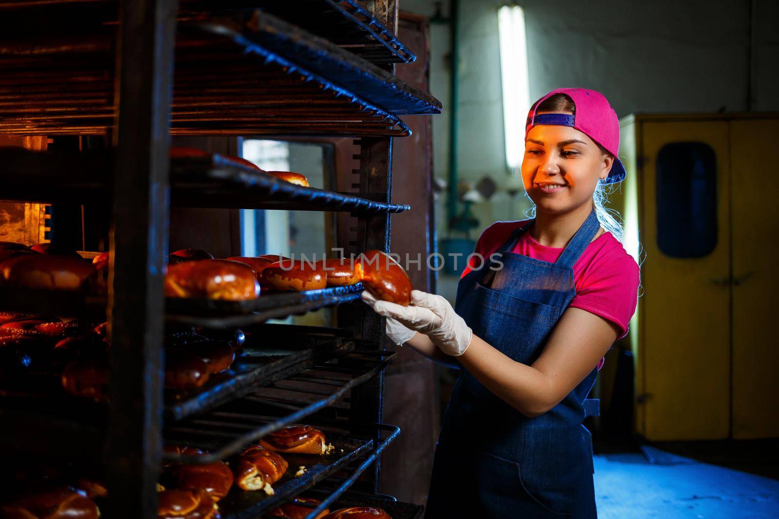 Girl baker holds fresh pies in the hands of the bakery. She is wearing a denim frock and a cap. Production of bakery products. Rack with hot crispy pastries.