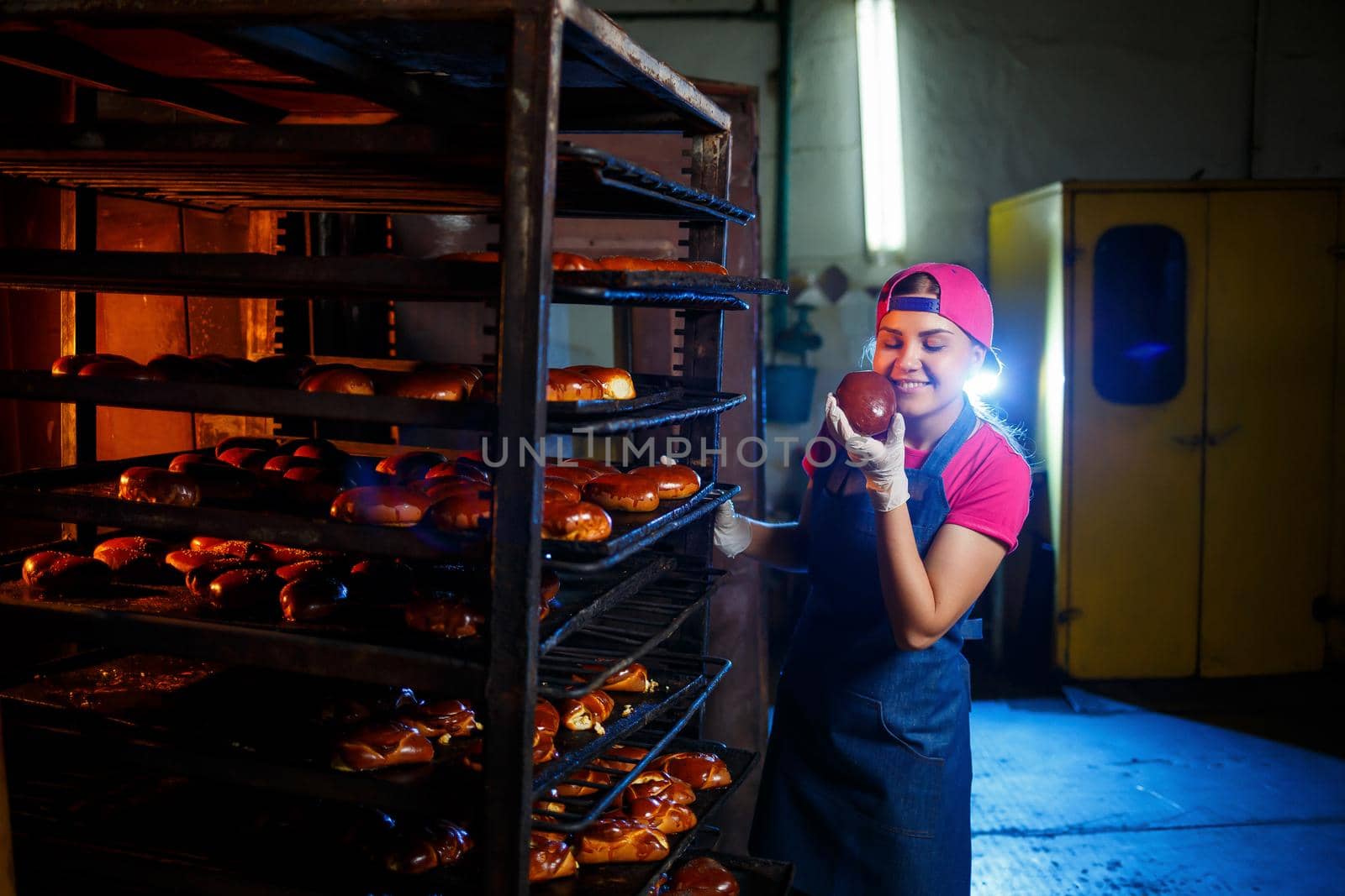 Girl baker holds fresh pies in the hands of the bakery. She is wearing a denim frock and a cap. Production of bakery products. Rack with hot crispy pastries.