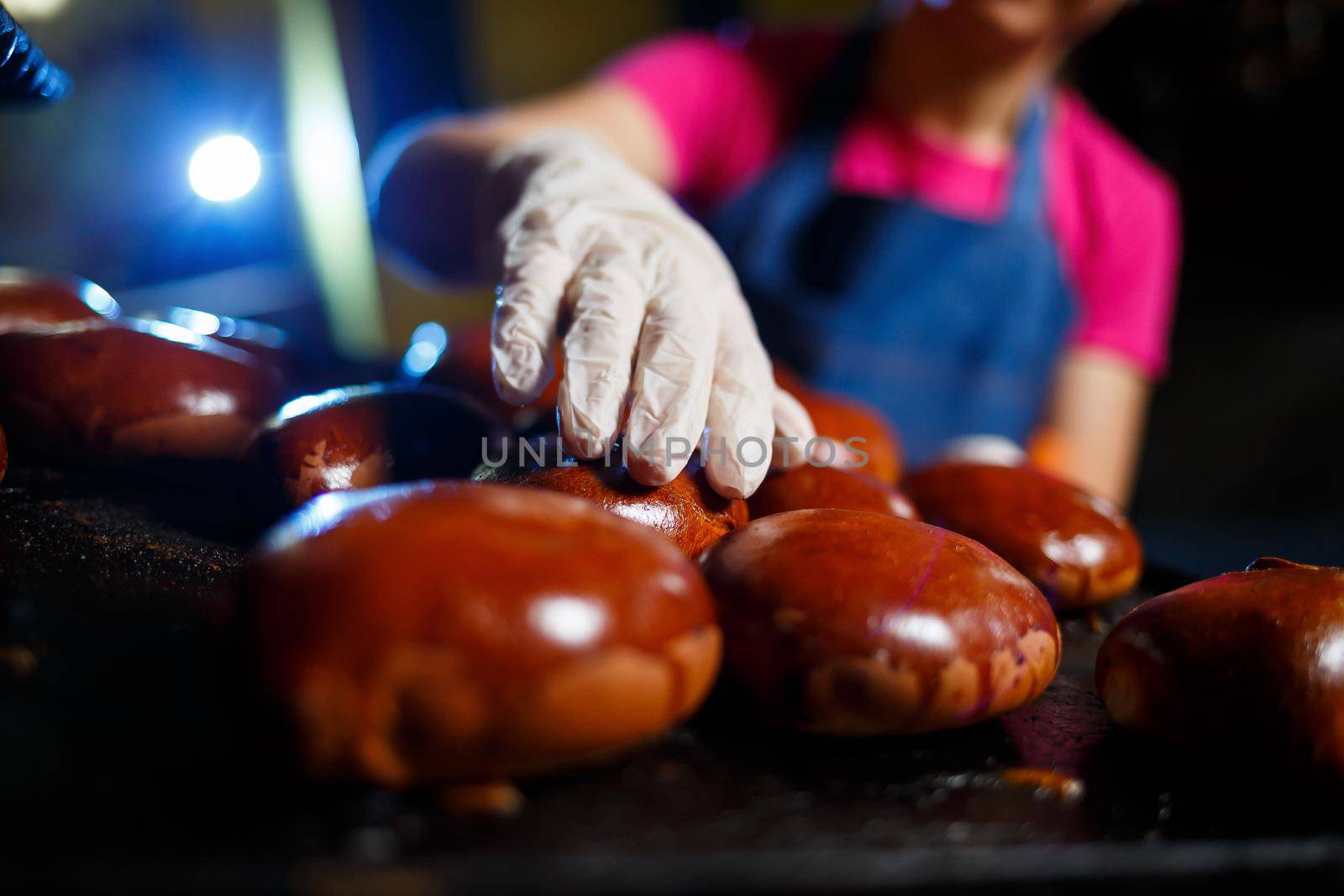 Girl baker holds a tray with hot pastries in the bakery. She is wearing a denim frock and a cap. Production of bakery products. Rack with fresh crispy pastries.