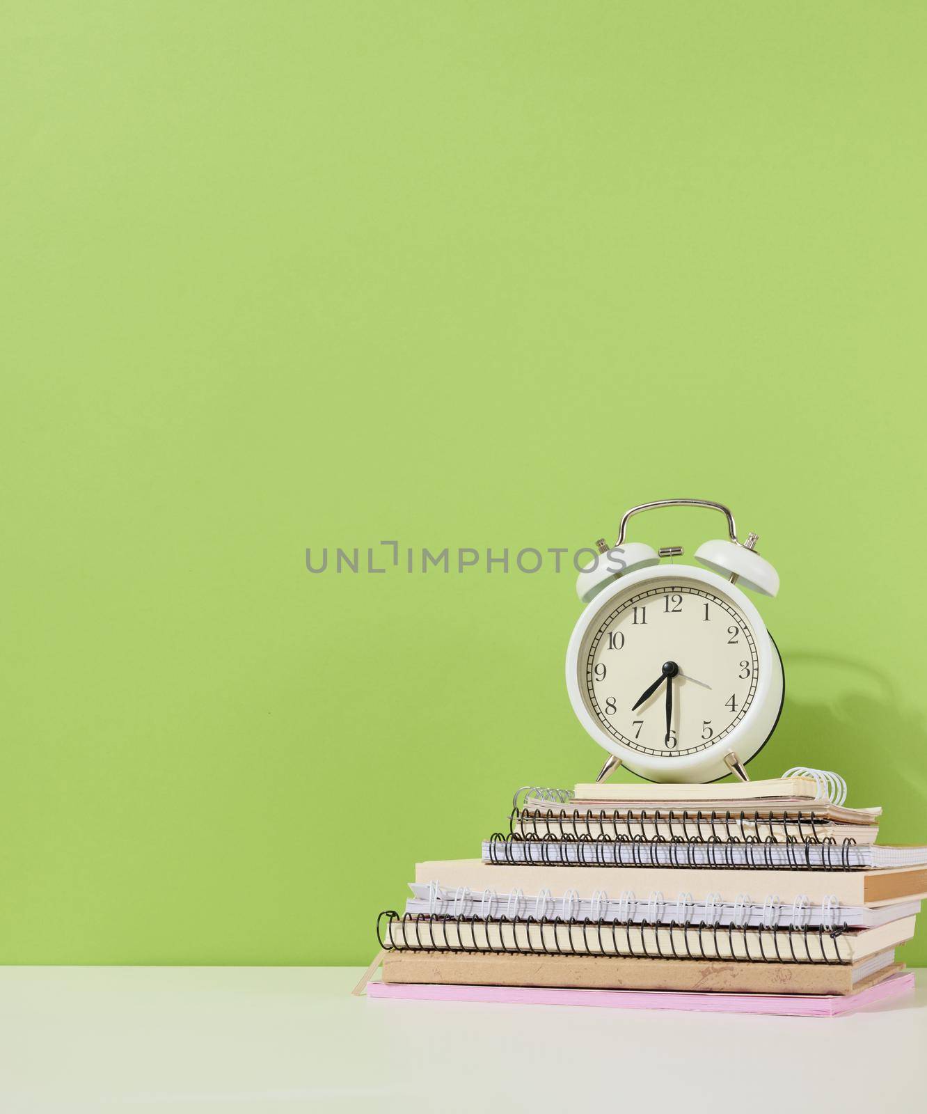 Round alarm clock stands on a stack of notebooks, green background. Back to school