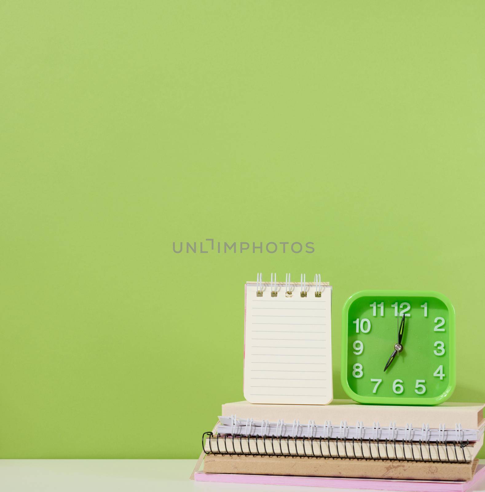 An open notepad and a green alarm clock stands on a stack of notebooks, a green background. Back to school