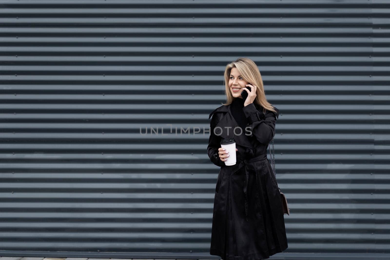modern businesswoman with a cup of coffee in her hands in business clothes speaks on the phone against the background of a gray textured wall.