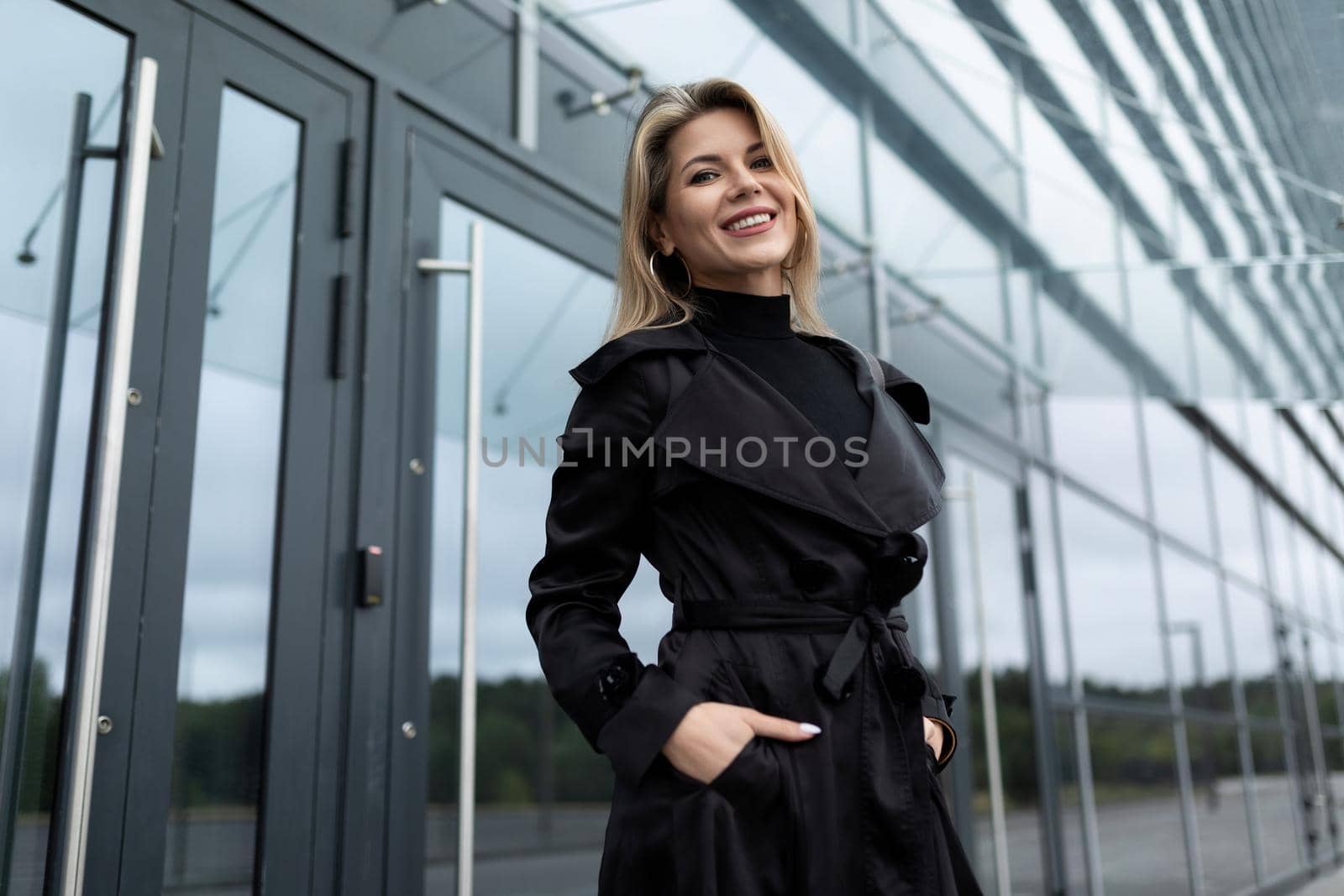 charming businesswoman in a black business coat at the door to the office building looking at the camera with a smile.