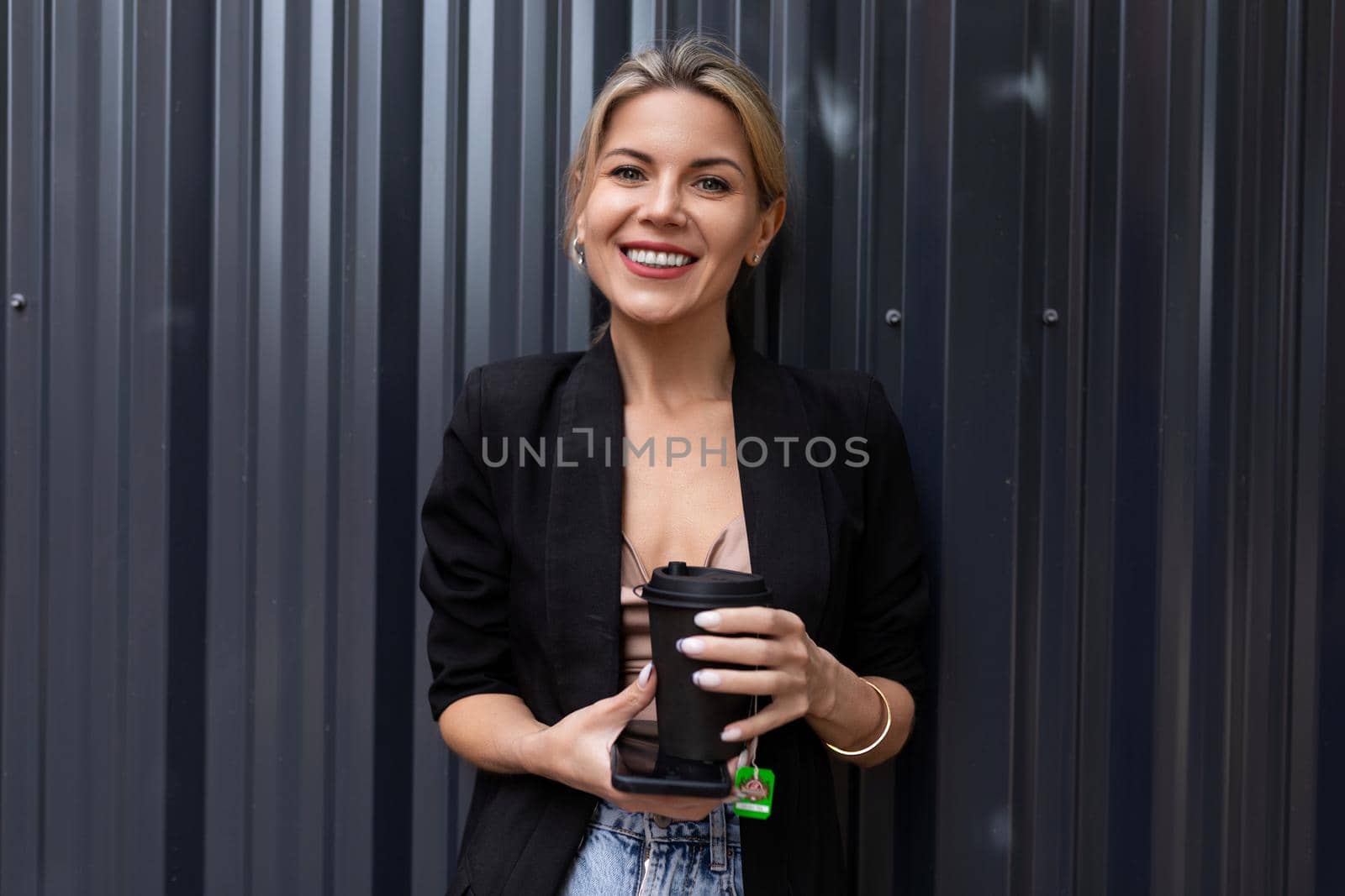 successful woman office employee with a mobile phone and a cup of tea on the background of a gray wall looks at the camera with a smile..
