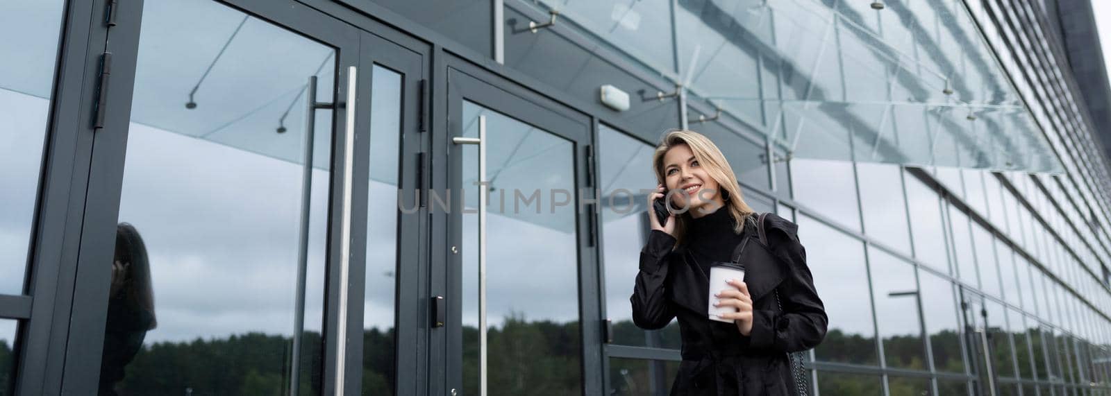 young woman businessman talking on the phone with a glass of coffee on the background of an office building by TRMK