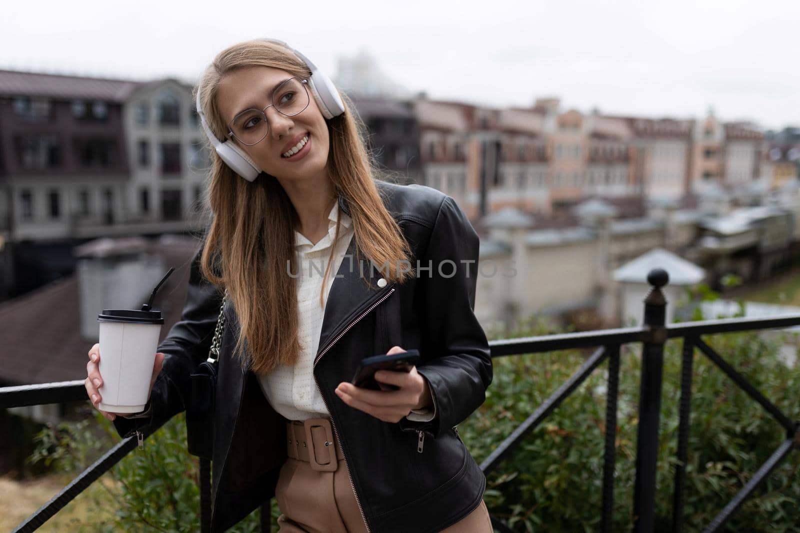 portrait of a cheerful and happy young woman with a mobile phone and a cup of coffee in her hands listening to music through over-ear headphones with a smile on her face.