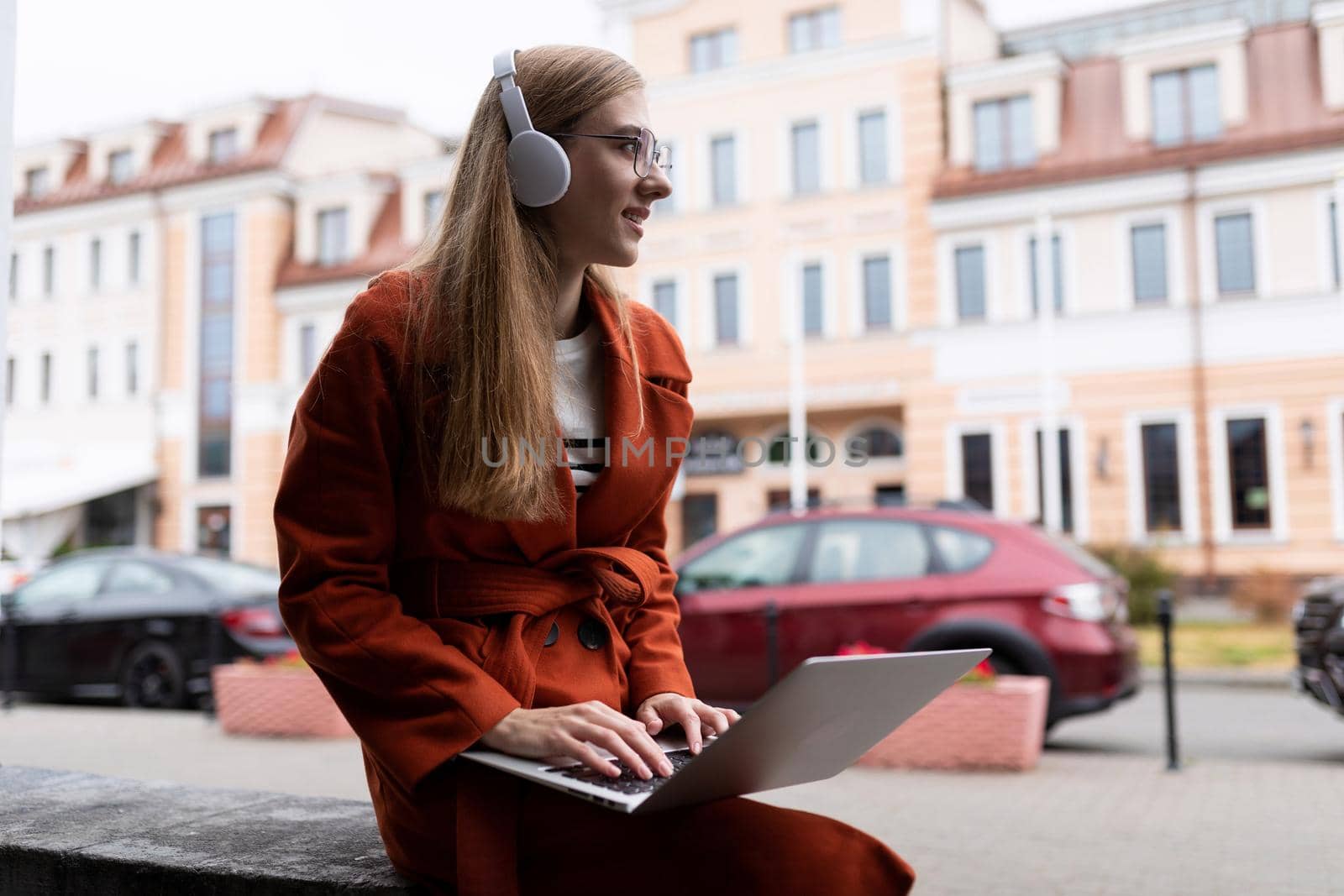 a young girl student in an autumn coat works on a laptop online and listens to music through headphones in the city.