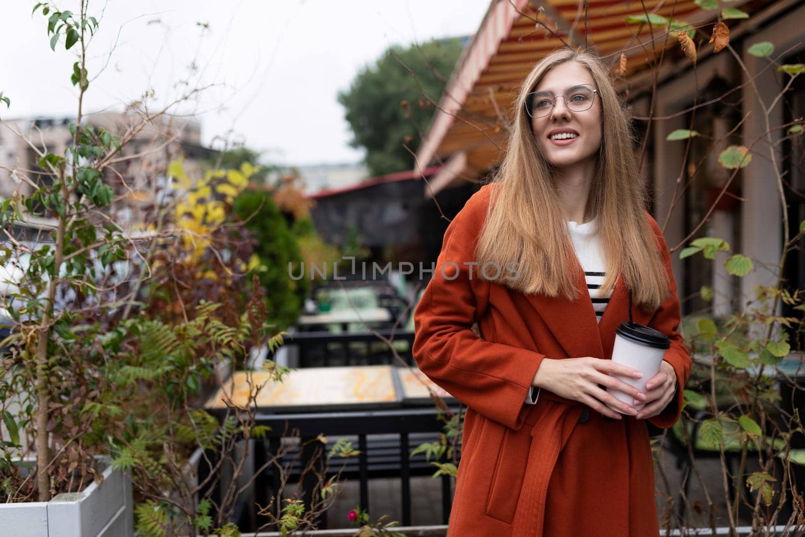 a young student with a cup of coffee in her hands next to a street cafe with a smile on her face.