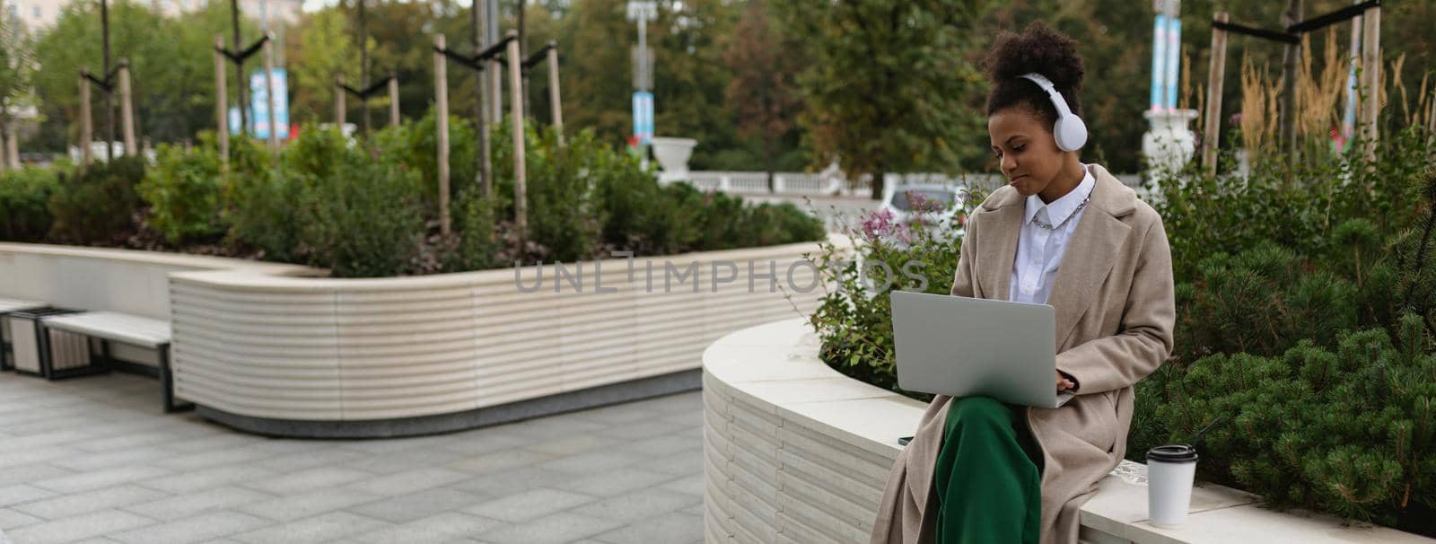 young black woman working online on a laptop in headphones with a cup of coffee in a city park with a smile on her face by TRMK