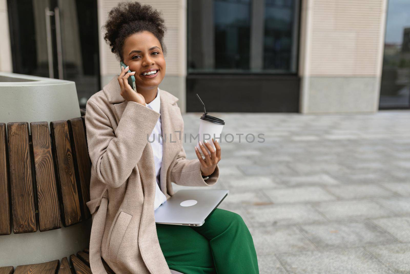 young african american woman sitting near the business center during lunch break with a laptop and a glass of coffee speaks with a smile on the phone.