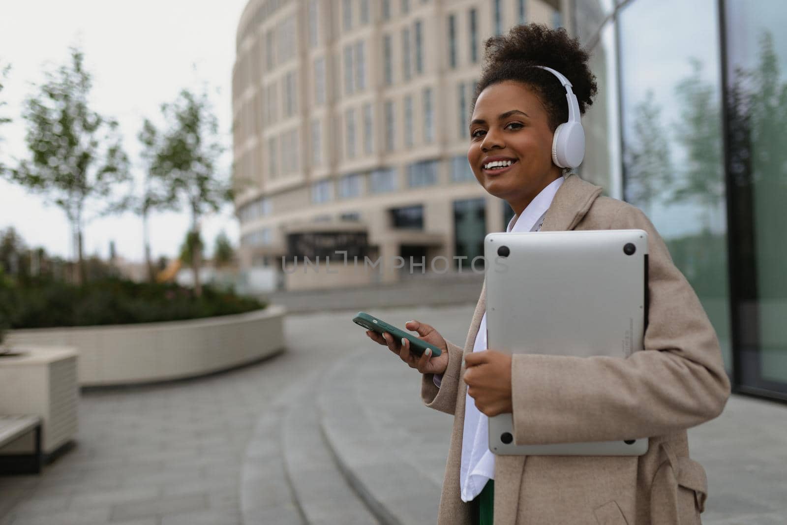 a young American woman with a wide smile walks in headphones with a mobile phone and a laptop against the backdrop of a city building by TRMK
