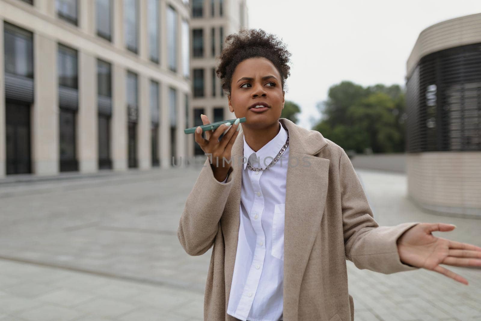 displeased african american woman talking on hands free mobile phone waving her hands against the backdrop of a business center.