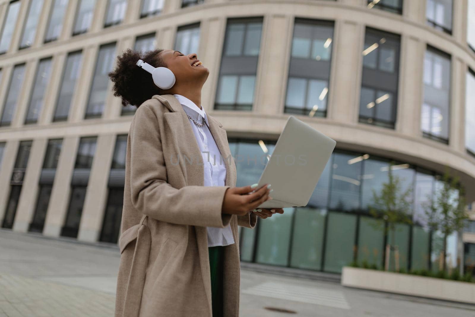 satisfied african american woman with a laptop in her hands and headphones emotionality laughs against the backdrop of an office building by TRMK