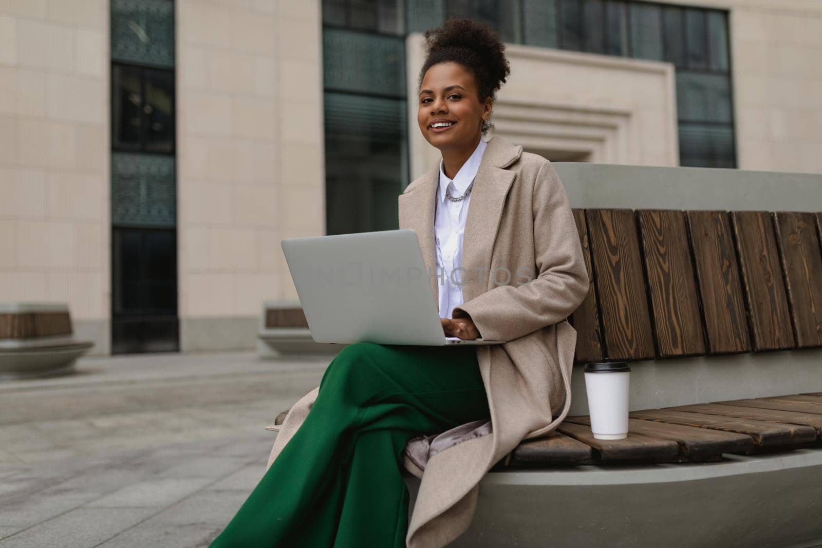 pretty successful young african american woman working on a laptop online sitting on a bench with a glass of coffee on the background of an office building by TRMK