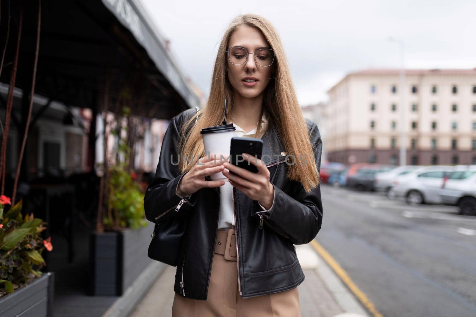 young woman typing a message on a mobile phone with a cup of coffee in her hands against the backdrop of the urban landscape next to a cafe.