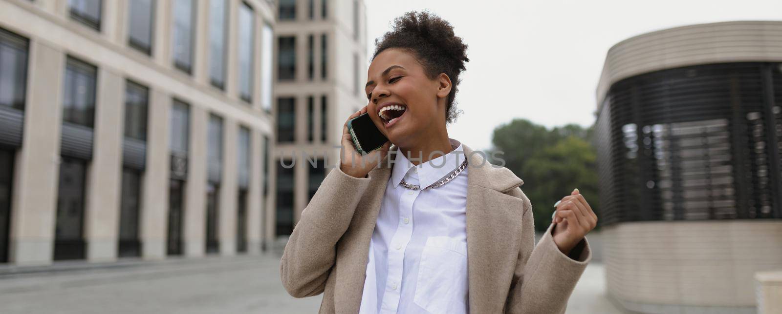 african american woman entrepreneur with a satisfied face and a smile speaks on a mobile phone after a successful transaction on the background of a business center by TRMK