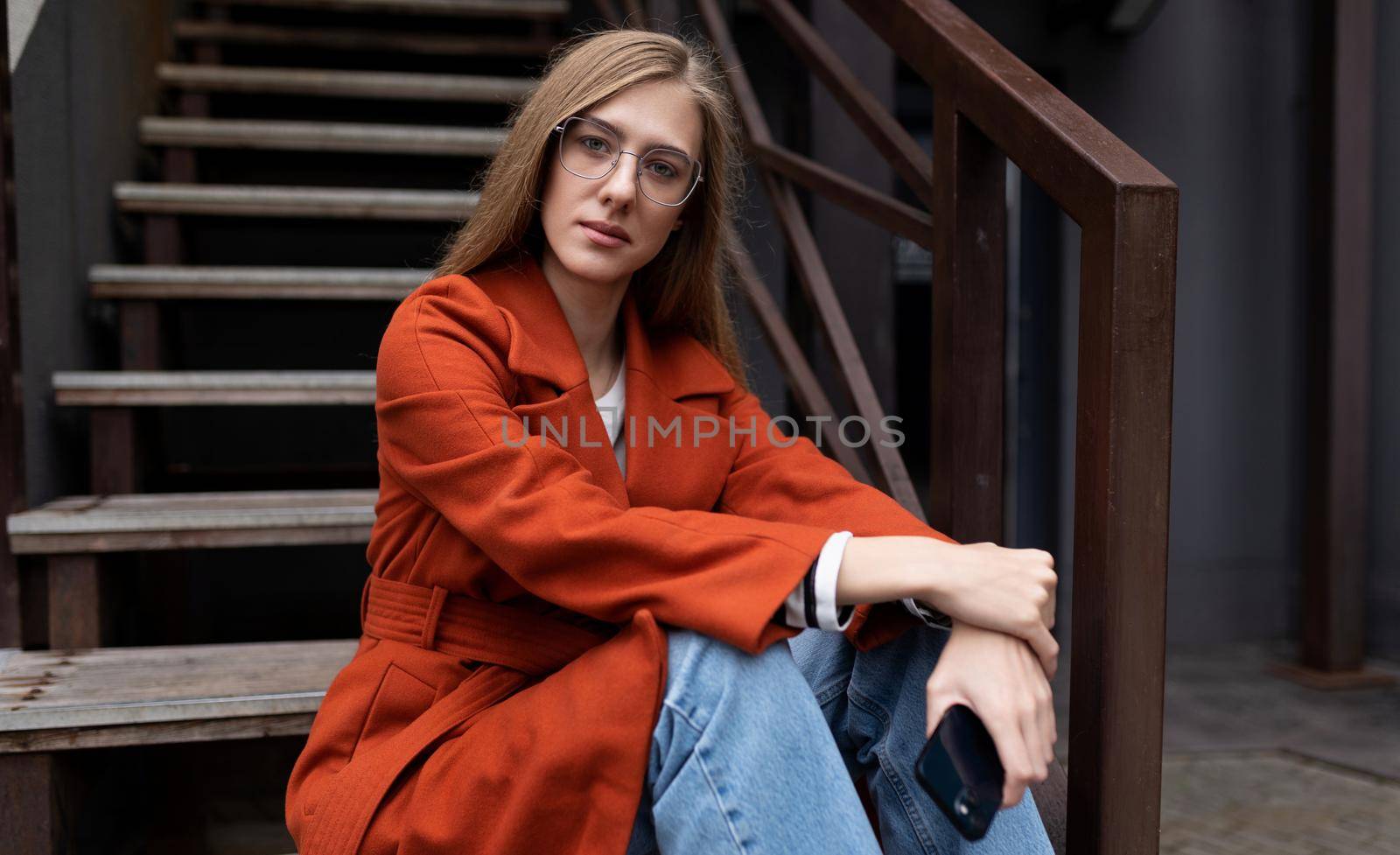 portrait of a pensive young woman in an autumn coat sitting on the stairs next to a cafe with a mobile phone in her hands.