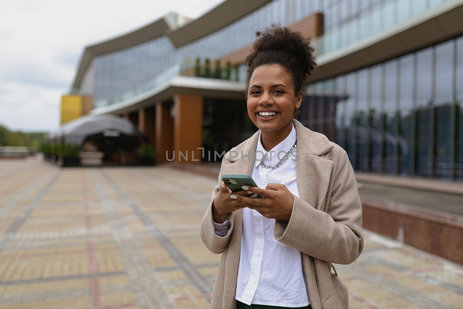 positive black young woman with a wide smile against the background of a modern city building with a mobile phone in her hands.