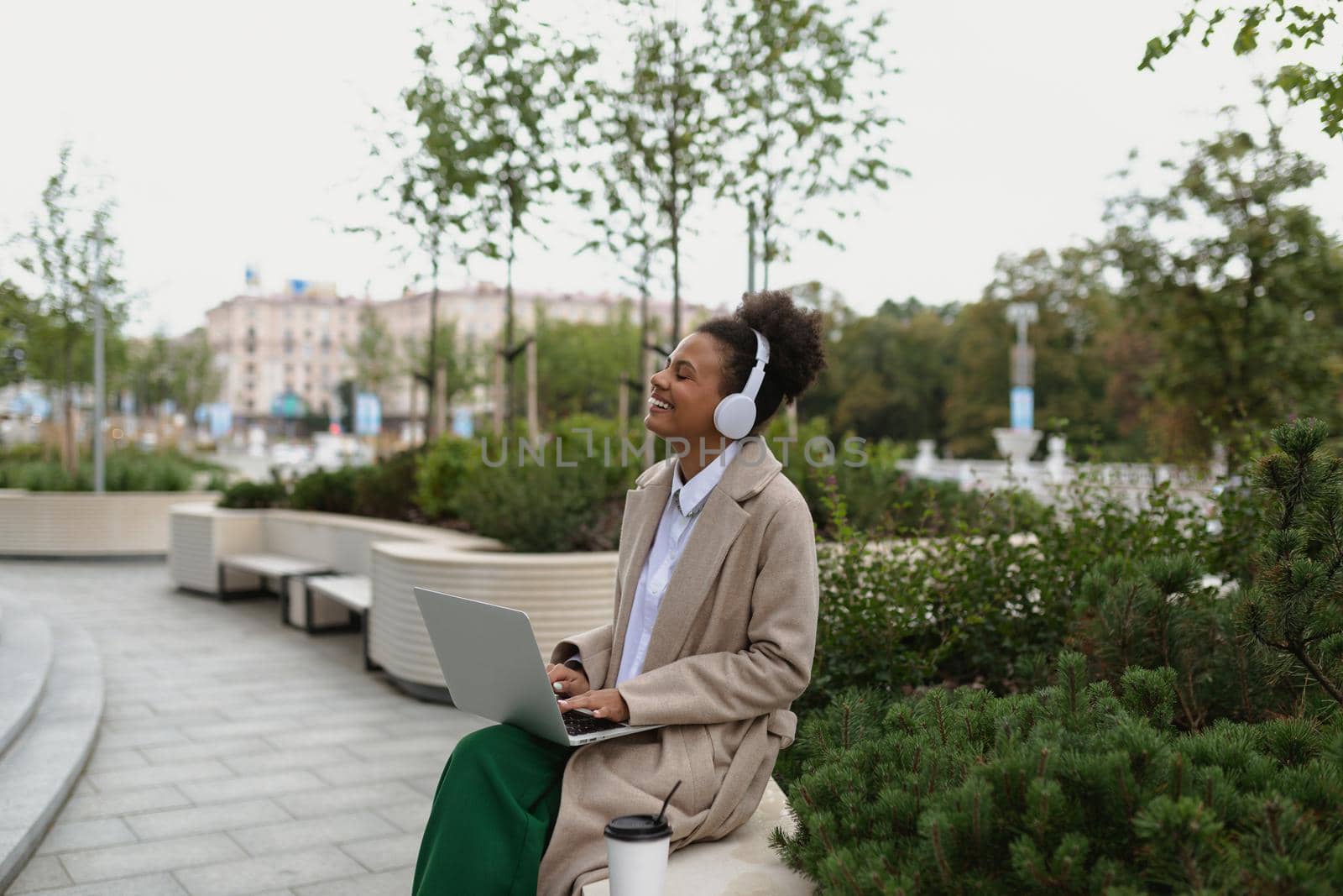 a young black student with a laptop sitting on a bench listening to music in headphones with pleasure closing her eyes.
