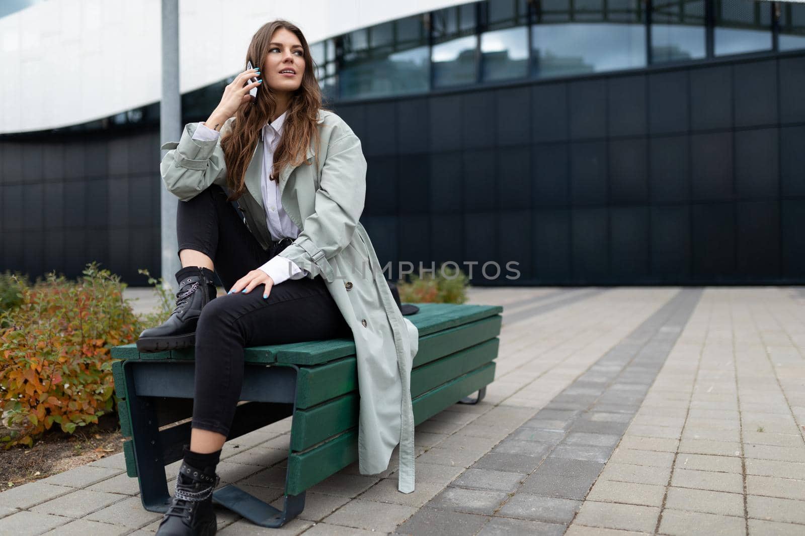 young woman talking on the phone sitting freely on a bench against the backdrop of an office building.