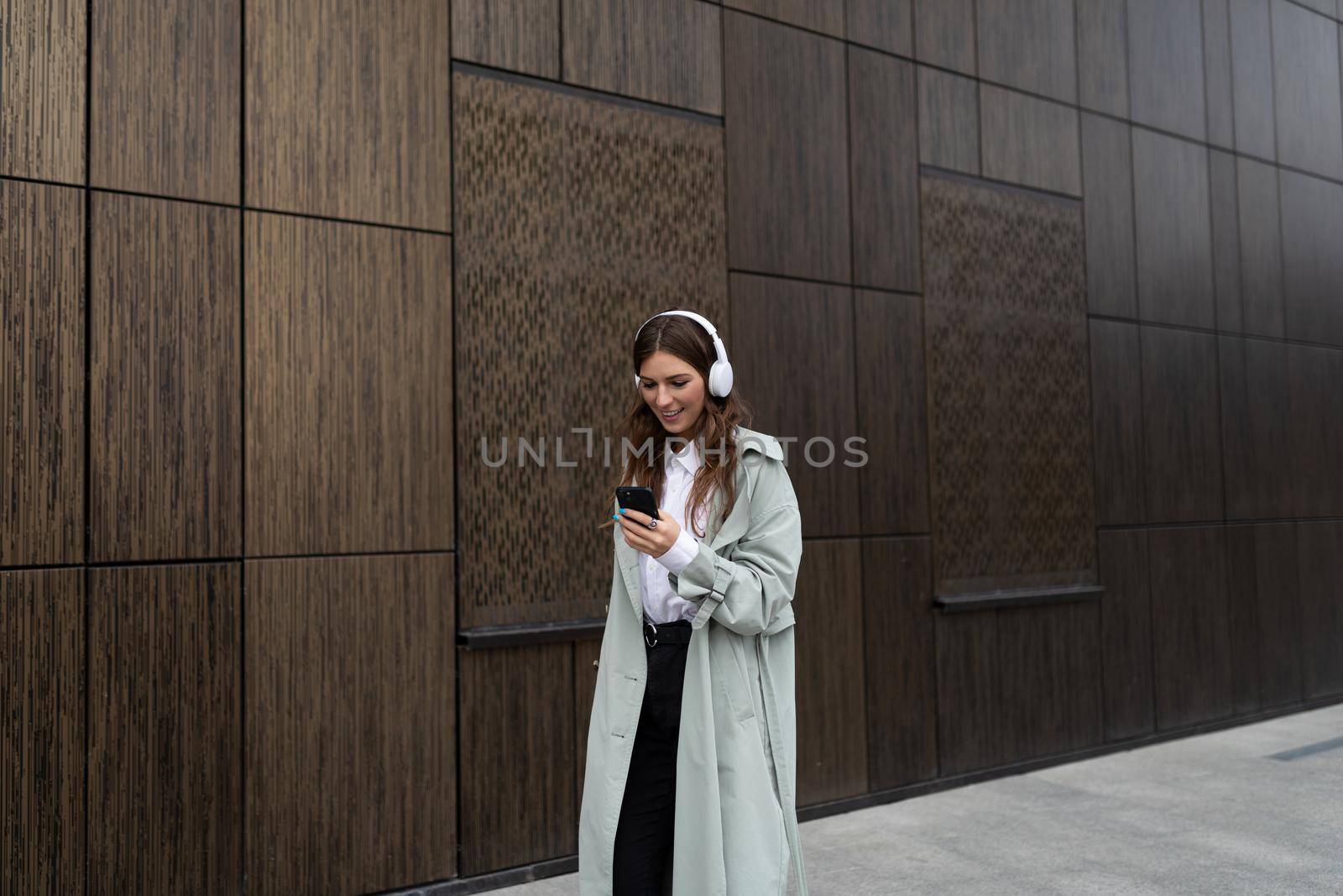 a young woman walks around the city against the background of the brown walls of a modern building in headphones listening to music.