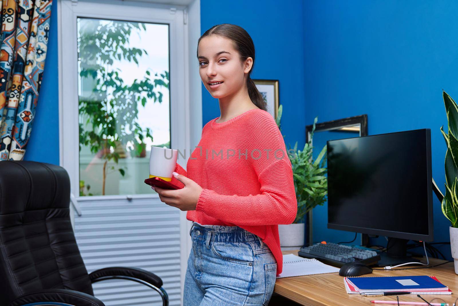 Teenage female at home near table with computer with smartphone and cup in hands by VH-studio