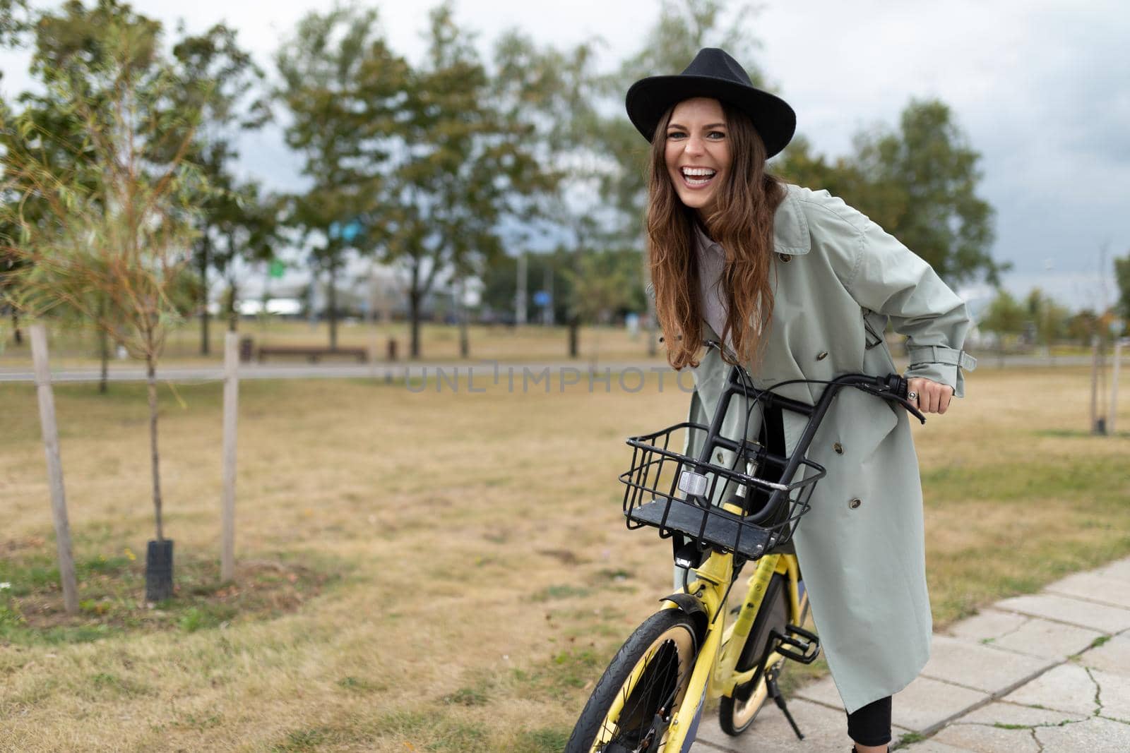 a young woman laughs while sitting on a bicycle while walking around the city in a black hat and a true gray coat.