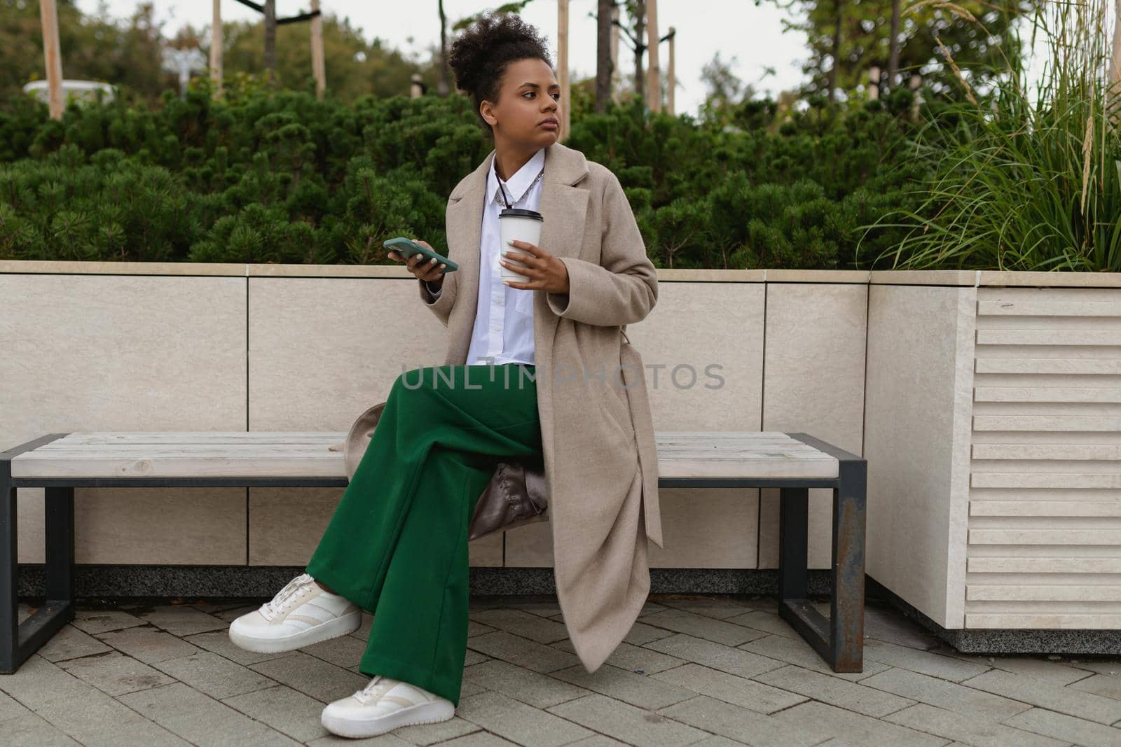 an african american woman sits on a bench in the park with a laptop and mobile phone looks thoughtfully into the distance.